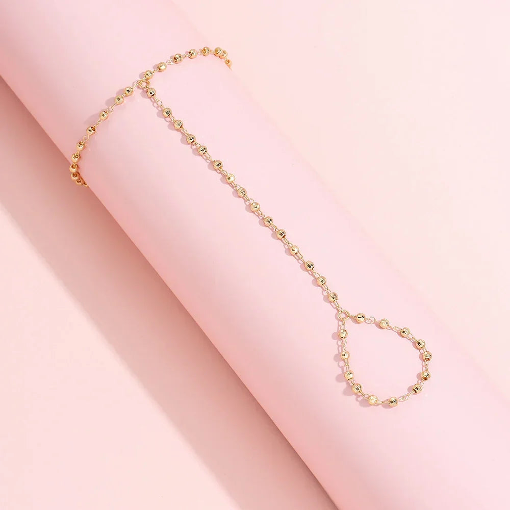 Elegant gold-plated faceted beads hand chain bracelet with finger ring displayed on pink background, emphasizing minimalist jewelry design.